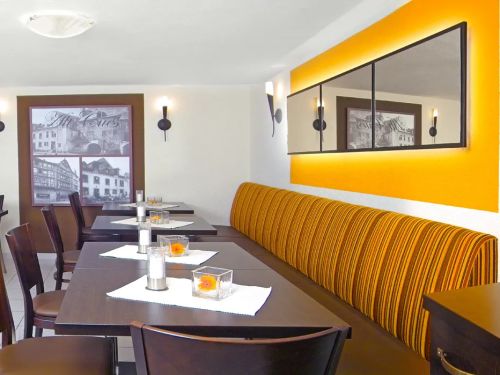 Gemütlicher Gastraum mit dunklen Holztischen Stühlen und gelb gestreifter Sitzbank an der Wand