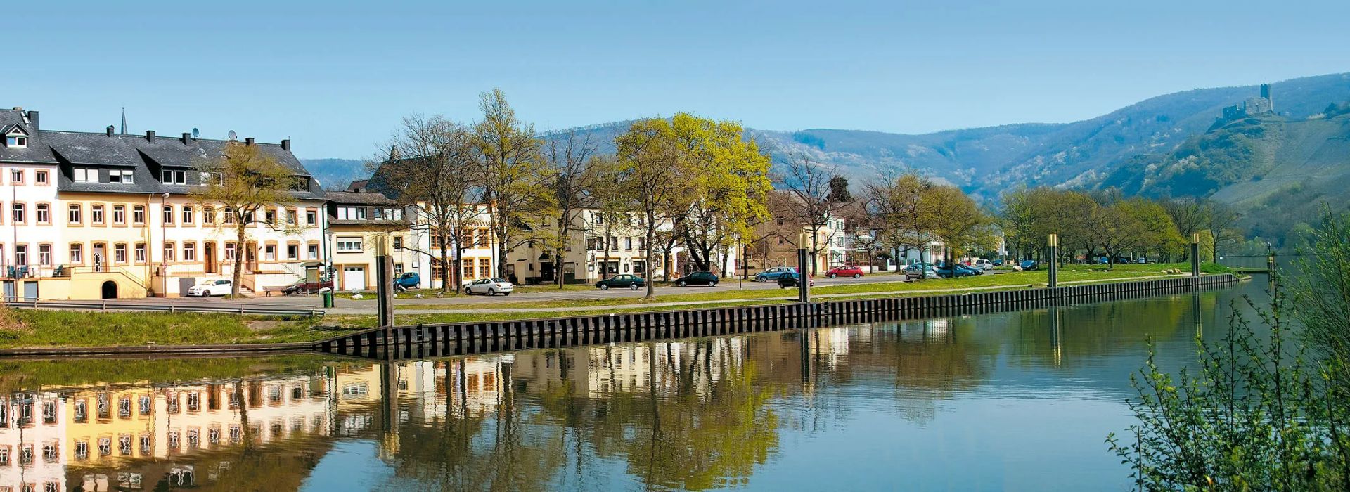 Häuser an einem Fluss bei sonnigem Wetter mit Bergen im Hintergrund und Spiegelung im Wasser